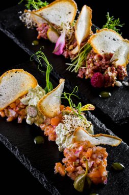 Buğday ekmekli tartar ve somonlu tartar, mercimek mikroyeşil lahanası, kapari, ahududu ve yenilebilir taç yaprakları. Yemekler siyah bir tabakta, tabaklar siyah bir zeminde..