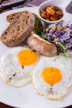Fried eggs from two eggs with grilled pork sausage, purple cabbage salad with corn. Lies on a white plate. On the plate are rye ciabatta toast and a sauce bowl with pickled beans. The plate stands on a wooden background, a knife and a fork lie next t