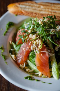 Somonlu salata, susam tohumu, roka, salatalık, salatalık, marul ve pesto sosu ahşap bir arka planda ekmek ile bir tabakta.