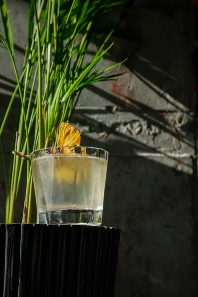 Cocktail with ice, dried pineapple and olive on a skewer in a glass that stands in a vase with herbs
