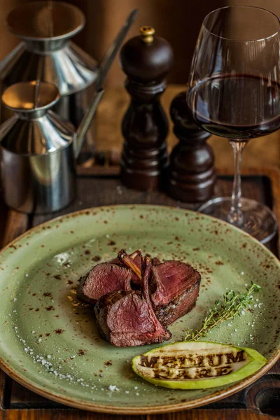 Rack of veal, medium rare, sliced in portions. Next to it is a sprig of thyme and a slice of zucchini with a roast signet. The food is on a green ceramic plate. The plate is on a wooden board, next to it are metal dishes for oil and spices, a glass o
