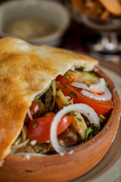 Domates, kabak, soğan, patlıcan ve hamur kapağıyla pişirilmiş havuç. Sebzeler odun ateşinde pişiriliyor. Tencere seramik tabakta. Yakınlarda, Gürcistan baharatlı seramik kaseler, bir bardak kırmızı şarap ve metal kâse wi var.