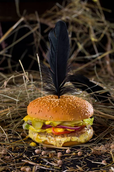 Burger with sesame bun and black feather stuck on top, cutlet, yellow sauce, lettuce, cheese and tomatoes, fresh onions and pickled cucumbers on the table with chickpeas and hay on a black background. Vertical orientation