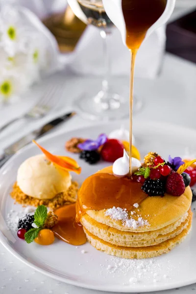 Ahududu, ahududu, kırmızı frenk üzümü, kurabiye kırıntıları ve vanilyalı dondurma içeren beyaz bir tabakta duran seramik bir sos teknesinden alınmış krep. Beyaz, seramik bir tabak beyaz bir masa örtüsü üzerinde duruyor, bir bardak beyaz şarap, bıçaklı bir çatal