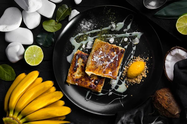 Casserole with condensed milk, powdered sugar, lemon and nut crumbs in a plate on the table with bananas, lime, coconut, white stones, rose leaves, and a black cloth