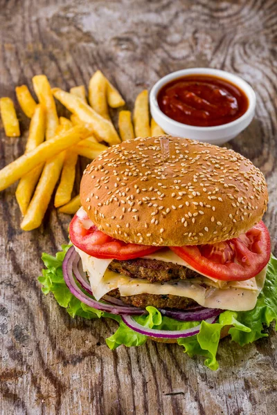 Burger with sesame bun, lettuce, double meat cutlet, tomatoes, onions and cheese on a wooden table with fries and sauce. Vertical orientation