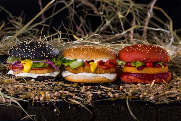 Three burgers with different fillings on a table with chickpeas and a sprinkle of hay on a black background. Horizontal orientation
