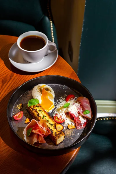 A slice of cheesecake with thyme, prosciutto, parmesan, basil, tomatoes, garlic and a boiled egg in a plate on the table with a cup of coffee