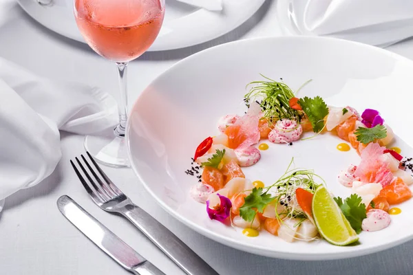 Salmon tartar and scallop tartar, next to it is Japanese mayonnaise with tobiko caviar, cilantro leaves, pickled cabbage, onion microgreen, chili slices and a drop of mango sauce. The food is in a white, deep, round plate. The plate is on a white tab