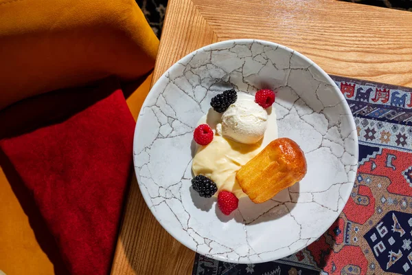 Savarin soaked in rum with vanilla ice cream and vanilla cream. Topped with raspberries and blackberries. The dessert lies in a round, deep plate with a pattern in the form of cracks. The plate stands on a wooden table with a traditional oriental blu