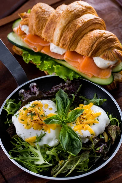Eggs benedict with iceberg lettuce and a sprig of basil in a small black frying pan, next to a croissant with salmon, mozzarella, lettuce and cucumber. Food stands on a wooden background.