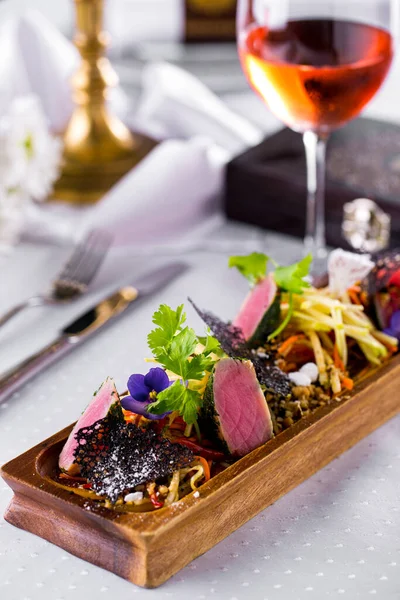 Tuna with parsley, black lace chip, violet flower and herbs on a wooden block with a glass of white wine, cutlery on a white tablecloth. Vertical orientation 