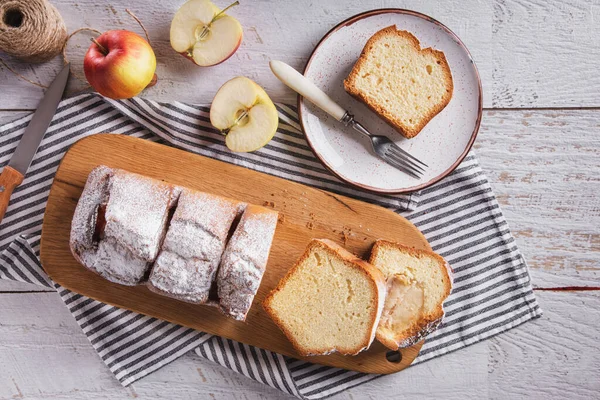 Sweet white bread sliced into pieces with powdered sugar, apples lying on a wooden board with a towel and a fork on the table