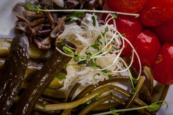 Domates, salatalık, kuşkonmaz, lahana ve mantardan yapılmış çeşitli sebze turşusu. Üstünde mercimek mikroyeşil lahanaları var. Yemek beyaz, oval bir tabakta. Tabak ahşap bir zeminde duruyor, yanında iki çatal var, bir dekan var.