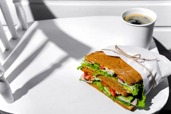 Sandwich in white paper with ham, lettuce, tomatoes, cucumbers and sauce with cappuccino with a paper cup standing on a white chair against the background of a white wooden wall