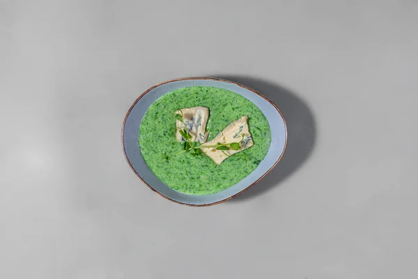 Creamy spinach soup topped with two slices of dorblu cheese and microgreen sprouts. Soup lies in a blue, ceramic bowl. The bowl stands on a gray paper background.