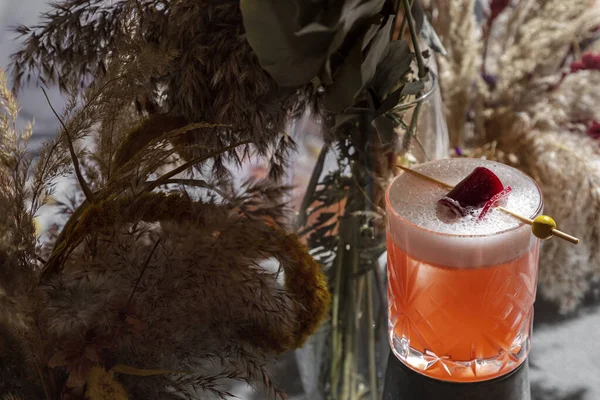 Cocktail with marmalade and olive on a stick in a glass that stands on a stand against a background of dried flowers