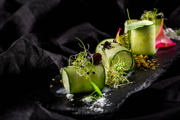 Kabak dürümleri, ince dilimlenmiş, baharatlı otlu tavşan ezmesine sarılmış. Üstünde soğan mikroyeşil lahanaları var. Ekmekler siyah bir tabakta duruyor. Yanında da ezilmiş fındıklar ve yenilebilir yapraklar var. Tabak siyah, keten bir kumaşın üzerinde..