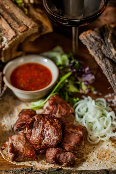 Grilled pork skewers, lying on pita bread, next to lettuce and arugula salad and sliced onion rings. A sauce-pot with adjika lies nearby, Food and sauce are on a wooden board. The board stands on a wooden background with acacia logs. A glass of red w