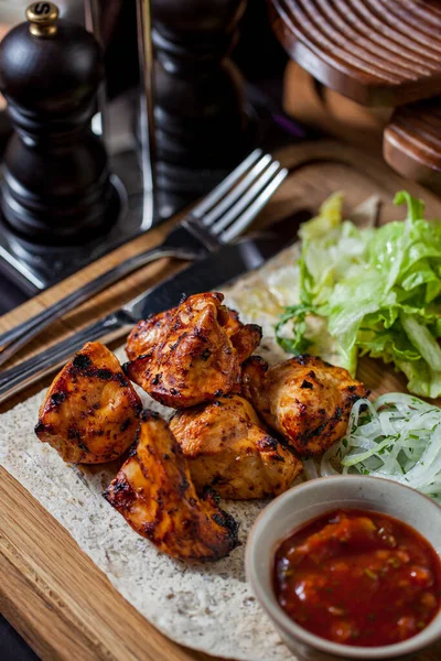 Chicken kebab on pita bread, next to it lies a cabbage salad, a bowl with vegetable sauce. The food is on a wooden board with a fork and knife nearby. The board is on a wooden table, next to it there are a pepper shaker and a salt shaker.