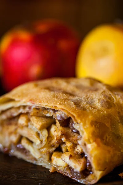 Strudel with apples, raisins, cinnamon and walnuts, baked in a thin, shortcrust pastry. The strudel lies on a wooden surface. There are two whole apples behind, one red and one yellow. Close-up.