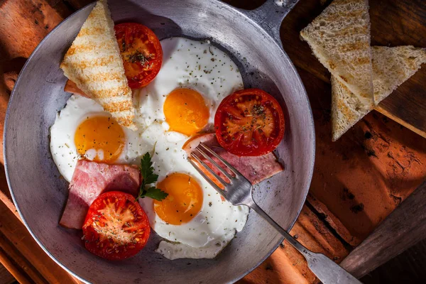 Fried eggs from three eggs with ham, fried tomatoes and toast. A fork is on top. Scrambled eggs are in a metal frying pan. A frying pan rests on a ceramic tile, next to it are two grilled toast.
