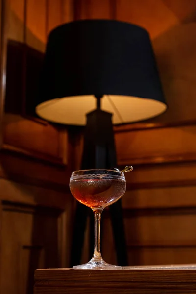Red vermouth with olive on a skewer. Vermouth is poured into a crystal glass with a pattern on a long stem. The glass is on the edge of the wooden table. Behind, in the background, you can see a chandelier with a black floor lamp, from which a lamp s