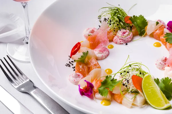 Salmon tartar and scallop tartar, next to it is Japanese mayonnaise with tobiko caviar, cilantro leaves, pickled cabbage, onion microgreen, chili slices and a drop of mango sauce. The food is in a white, deep, round plate. The plate is on a white tab
