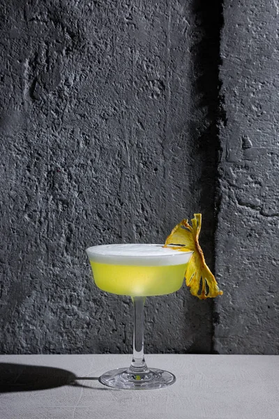 A cocktail with ice, froth and dried pineapple in a glass that stands on the table against the background of a stone wall