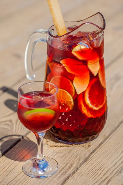 Red wine sangria decanter with apple, orange, lemon, lime wedges, raspberries, grapes and ice. There is a glass of ice, raspberries and sangria nearby. Stands on a wooden background illuminated by sunlight.