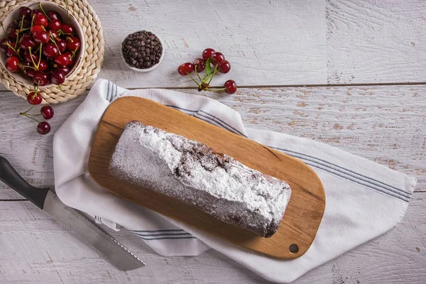 Sweet white bread with powdered sugar, cherries and chocolate droplets lying on a wooden board with a towel and a knife on the table