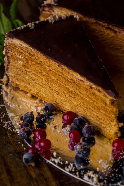 A piece of honey cake in chocolate glaze with currants, mint and cranberries on a wooden table. Vertical orientation