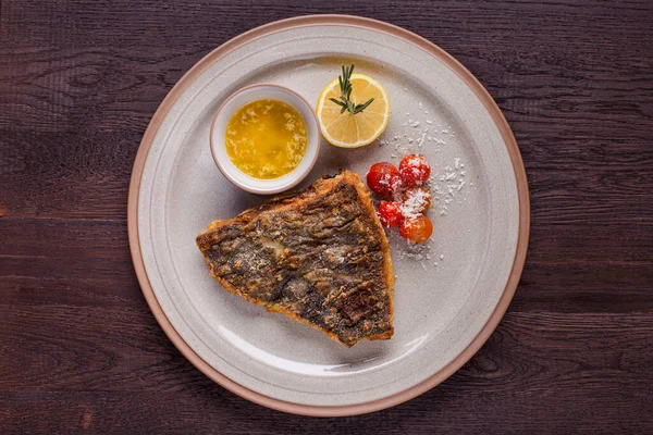 Fried fish steak with sauce, rosemary, half lemon, parmesan syrup and cherry tomatoes on a round plate and a wooden table. Horizontal orientation