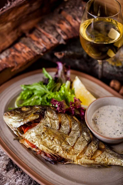 Fried and grilled dorado with white sauce with parsley, a slice of lemon, mung bean salad, arugula on an oval plate with a glass of wine. Vertical orientation 