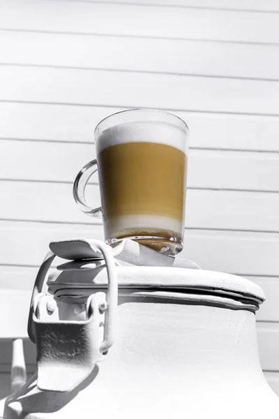 Glass of cappuccino with froth on a white can on a white chair against a background of a white wooden wall