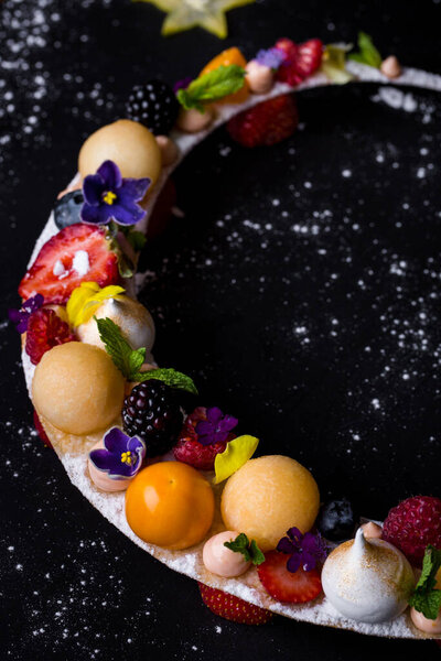 Dessert with beige balls, meringues, raspberries, blueberries, mint, flower petals, blackberries, strawberries, kumquat which lies on powdered sugar in the form of a crescent on a black background