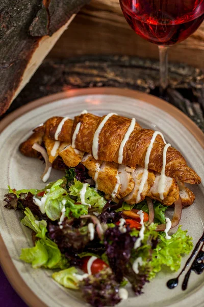 Croissant with grilled chicken fillet, lettuce, tomatoes and garlic sauce. Next to it is a mix of green and dark salad with cherry tomatoes and garlic sauce. They lie on a ceramic plate. The plate is on a wooden table, there is wood for the fireplace