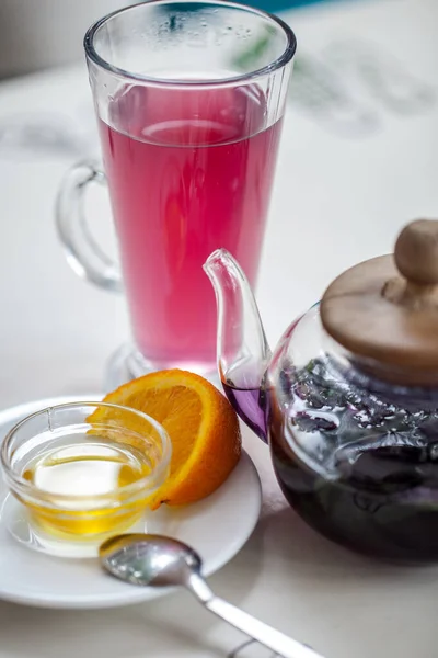 A hot drink made from purple basil in a clear glass glass. Next to the glass is a transparent glass teapot with a drink and basil leaves inside. Nearby lies a saucer with an orange slice and a small one. transparent bowl with honey. The lid of the te