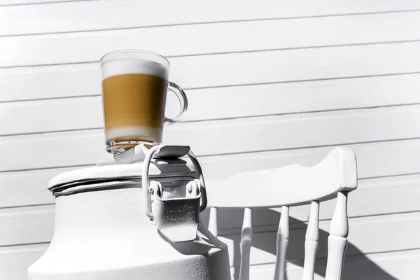 Glass of cappuccino with froth on a white can on a white chair against a background of a white wooden wall