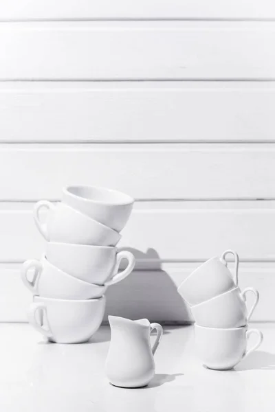 Cups for coffee and tea standing on top of each other and jug for cream on white table and white wood background