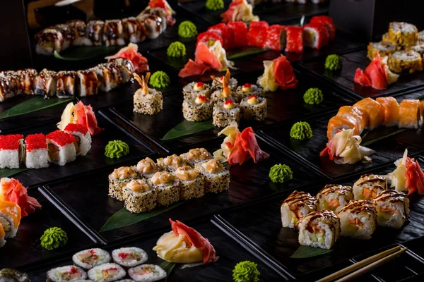 Different types of rolls with tuna, shrimp, scallops, salmon, crab, eel, tobiko caviar, rice, cucumbers, butter, sauce, sesame seeds, avocado, nori and Philadelphia cheese on black plates with wasabi, pickled ginger and banana leaves on a dark backgr