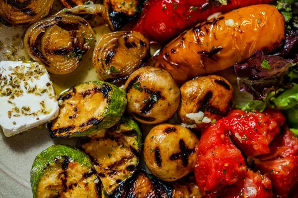 Grilled vegetables on the grill, red paprika, yellow paprika, zucchini, mushrooms, onion, tomato, with lettuce leaves, olive oil, feta cheese and herbs. They lie on a ceramic, oval plate, among the wooden logs.