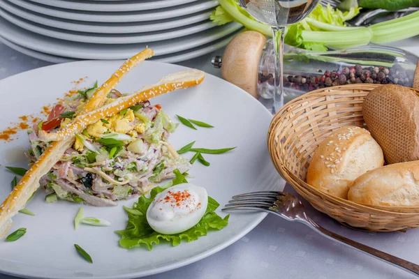 Salad with meat bread sticks, onions, boiled egg, arugula and salad in a plate on a light background with cutlery, baskets of bread and a glass of wine