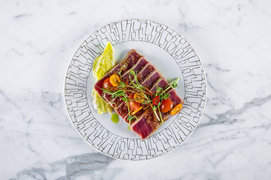 Fried pieces of tuna with young shoots of green peas, sauce, lettuce, tomatoes and kumquat on a white plate and on a light background. Horizontal orientation