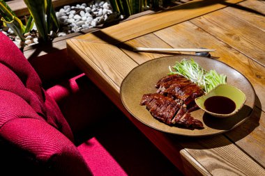 Fried chicken breast with leeks and cucumber in a plate with sauce in a bowl and chopsticks lying on the table and a red chair on the floor