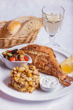 Grilled mackerel with sauce in, potatoes with porcini mushrooms, fried vegetables and lemon on a plate, a basket of bread and a glass of white wine 