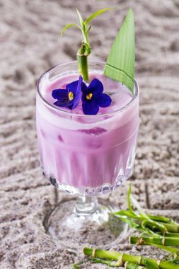 Purple cocktail with froth, ice, bamboo, violet and a leaf in a glass that stands on a sandy background