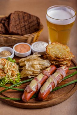 Peynirli domuz sosisi, bir bardak bira, yeşil soğan, lahana salatası, pita cipsi, patates kızartması, ahşap yuvarlak bir tahtada, kağıt bir arka planda.