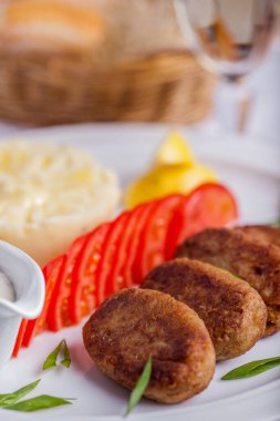 Four pork cutlets with finely chopped green onions, chopped tomato and mashed potatoes next to it. There is a gravy boat with mushroom sauce nearby. The food and sauce bowl are on a white, round plate and lemon wedges. The plate is on the table with 