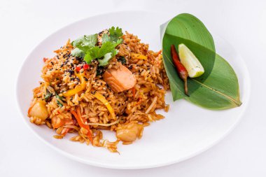 Rice wok with carrots, red paprika, yellow paprika, cilantro, salmon and scallop. On a plate nearby is a red chili pepper with a slice of lime, wrapped in a bamboo leaf. The food and decoration lie on a white, round plate. The plate stands on a white